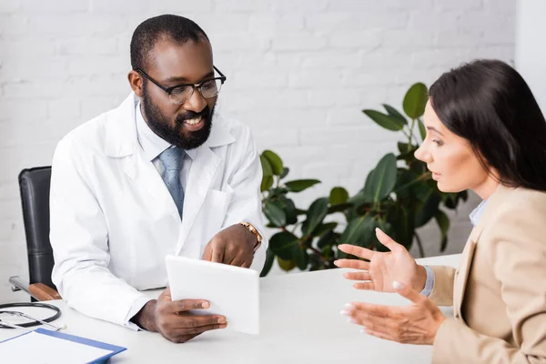 Selective focus of brunette woman talking to bearded african american doctor pointing with finger at digital tablet — Stock Photo