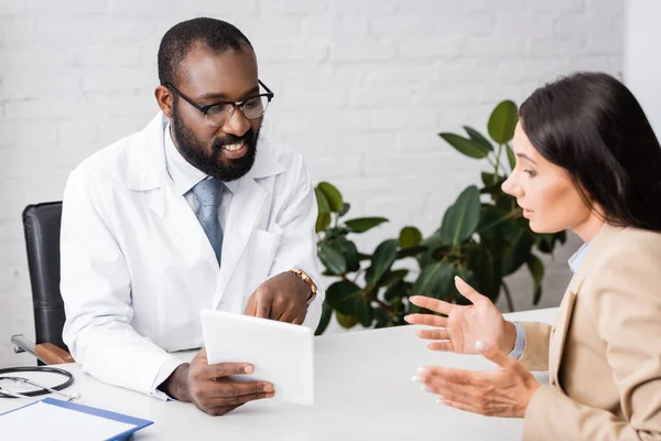 Selektiver Fokus einer brünetten Frau im Gespräch mit einem bärtigen afrikanisch-amerikanischen Arzt, der mit dem Finger auf ein digitales Tablet zeigt — Stockfoto