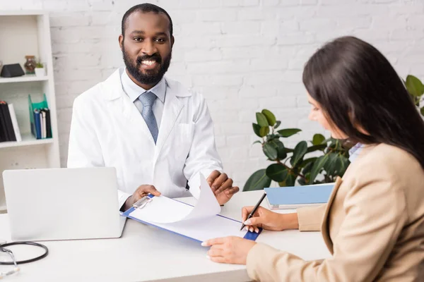 African american doctor holding clipboard with blank paper near brunette woman with pen — Stock Photo