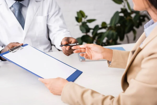 Partial view of woman taking pen and clipboard with blank paper from african american doctor — Stock Photo