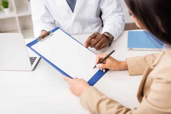 Ausgeschnittene Ansicht einer Frau, die Stift und Klemmbrett mit weißem Papier in der Nähe eines amerikanischen Arztes hält — Stockfoto