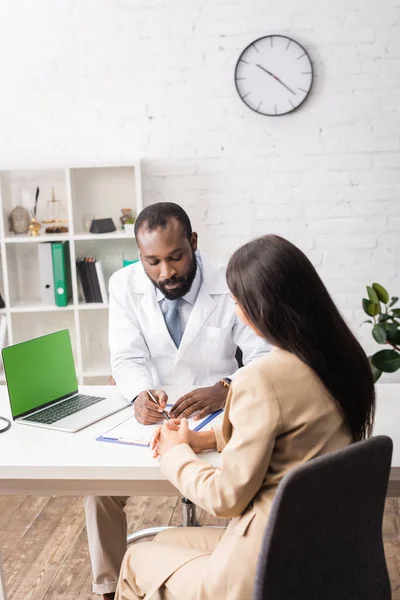 African american doctor writing prescrition on clipboard near brunette patient — Stock Photo