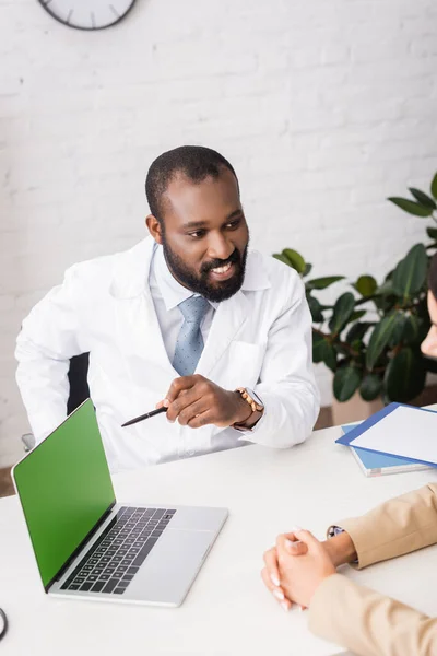 Selektiver Fokus des bärtigen afroamerikanischen Arztes, der mit Stift auf Laptop mit grünem Bildschirm in der Nähe des Patienten zeigt — Stockfoto