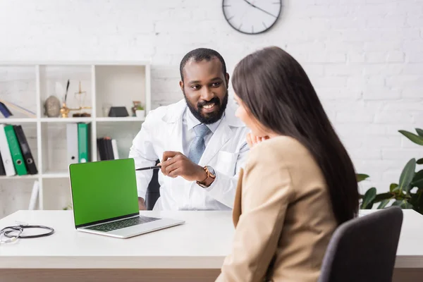 African american doctor pointing with pen at laptop with green screen near brunette woman — Stock Photo