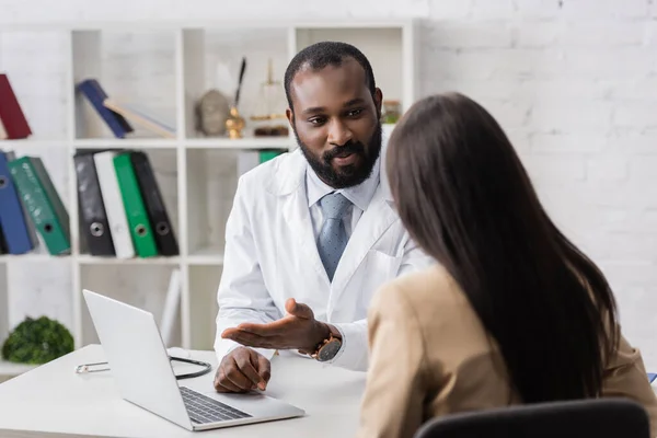 Visão traseira da mulher morena perto de barbudo Africano americano médico apontando com o dedo para laptop — Fotografia de Stock