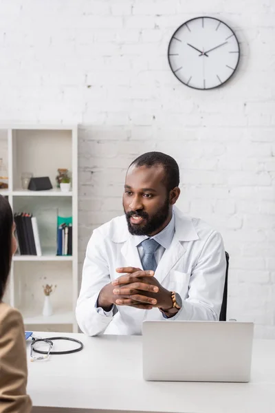 Foyer sélectif du médecin afro-américain assis avec les mains serrées en face du patient sur le lieu de travail — Photo de stock