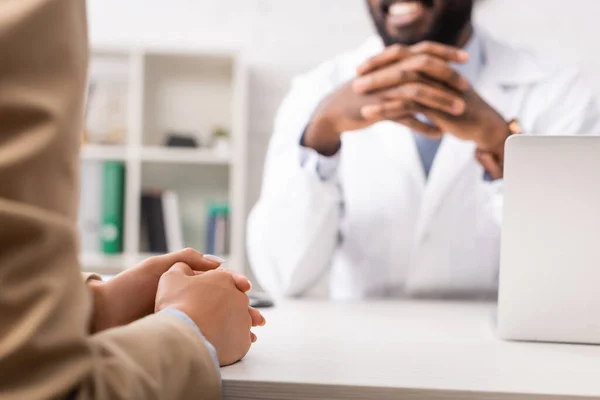 Vista recortada del médico afroamericano sentado con las manos apretadas cerca del paciente - foto de stock