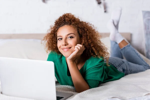 Selektiver Fokus einer jungen Frau, die in die Kamera blickt, während sie in der Nähe des Laptops auf dem Bett liegt — Stockfoto