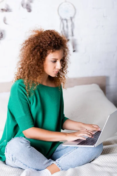 Selektiver Fokus einer jungen Frau mit Laptop, während sie mit überkreuzten Beinen auf dem Bett sitzt — Stockfoto