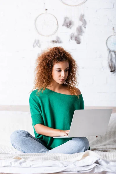 Selective focus of red haired woman sitting on bed and using laptop — Stock Photo