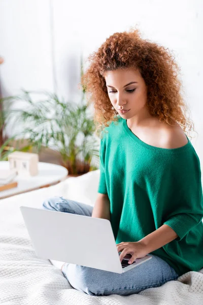 Selective focus of red haired woman in jeans using laptop on bed — Stock Photo