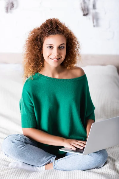 Curly woman in jeans looking at camera while using laptop on bed — Stock Photo