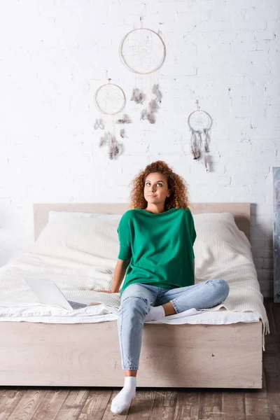Mulher sonhadora em jeans sentado na cama perto do laptop — Fotografia de Stock