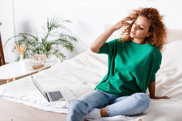 Mujer joven tocando el pelo cerca de la computadora portátil en la cama - foto de stock