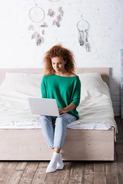 Червоне волосся жінка в джинсах використовує ноутбук, сидячи на ліжку — Stock Photo