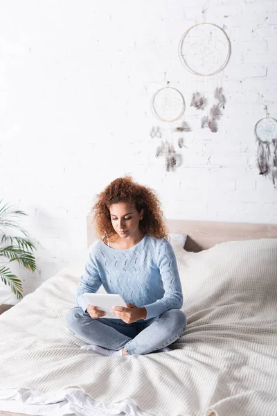 Young curly woman in sweater using digital tablet while sitting with crossed legs on bed — Stock Photo