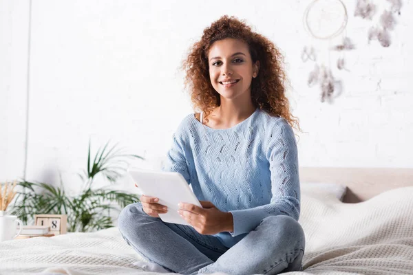 Rothaarige Frau im Pullover hält ein digitales Tablet in der Hand und blickt in die Kamera auf dem Bett — Stockfoto