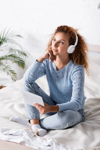 Selektiver Fokus einer verträumten Frau mit Kopfhörer, die ein digitales Tablet auf dem Bett hält — Stockfoto