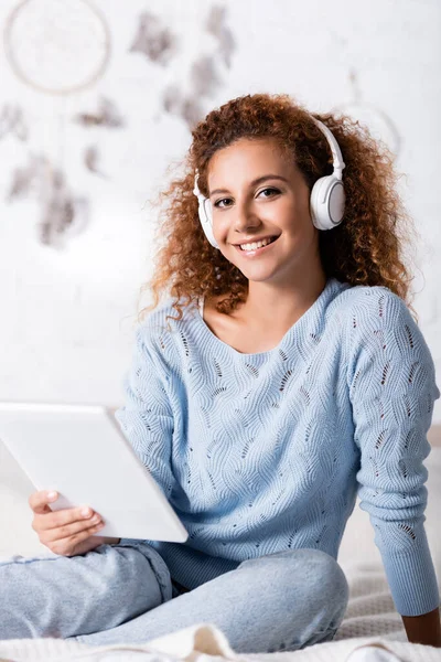 Selective focus of curly woman in headphones looking at camera while holding digital tablet on bed — Stock Photo