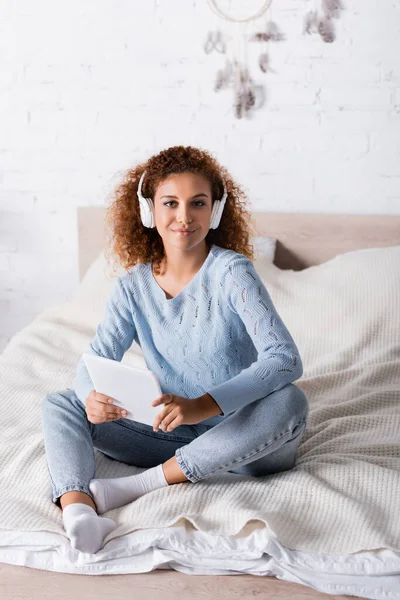 Lockige Frau mit Kopfhörer, digitales Tablet in der Hand und Kamera auf dem Bett — Stockfoto