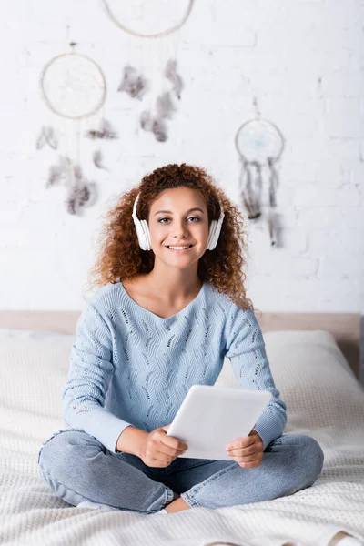Enfoque selectivo de la mujer en suéter escuchando música en auriculares y sosteniendo la tableta digital en el dormitorio - foto de stock