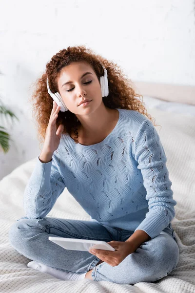 Femme rousse aux yeux fermés écoutant de la musique dans un casque et tenant une tablette numérique sur son lit — Photo de stock