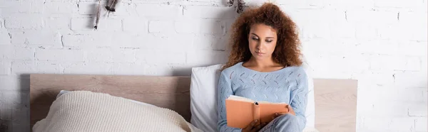 Foto panorámica de mujer joven en suéter libro de lectura en la cama - foto de stock