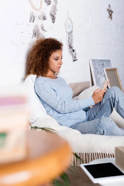 Selektiver Fokus der Frau beim Lesen von Büchern auf dem Bett in der Nähe des digitalen Tablets auf dem Couchtisch im Schlafzimmer — Stockfoto