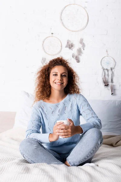 Selektiver Fokus einer jungen Frau, die in die Kamera blickt, während sie Tasse auf dem Bett hält — Stockfoto