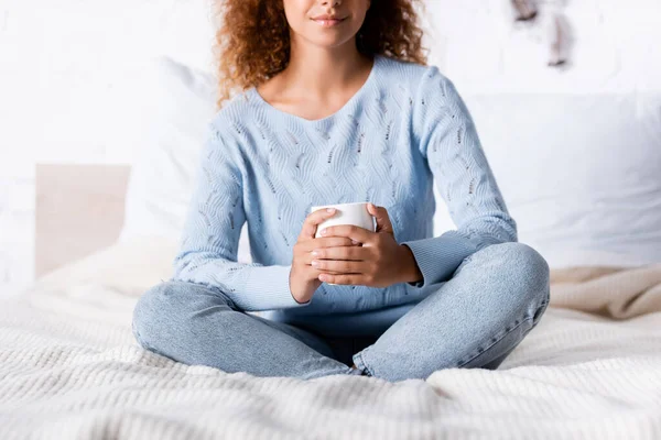Cropped view of curly woman holding cup on bed — Stock Photo
