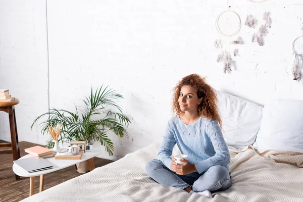 Junge Frau mit Tasse im Bett neben Laptop und Büchern auf Couchtisch — Stockfoto