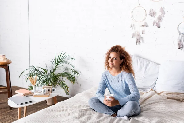 Young woman holding cup while sitting on bed near laptop and books on coffee table — Stock Photo