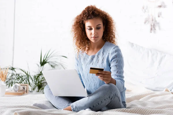 Вибірковий фокус кучерявої жінки за допомогою ноутбука та кредитної картки на ліжку — стокове фото