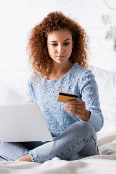 Вибірковий фокус кучерявої жінки, дивлячись на кредитну картку, тримаючи ноутбук у спальні — стокове фото