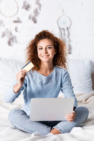Red haired woman looking at camera while holding credit card and laptop on bed — Stock Photo