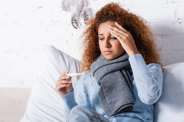 Selective focus of sick woman in scarf holding thermometer on bed — Stock Photo