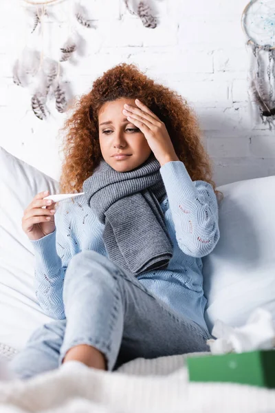 Вибірковий фокус втомленої жінки в шарфі, що тримає термометр на ліжку — стокове фото