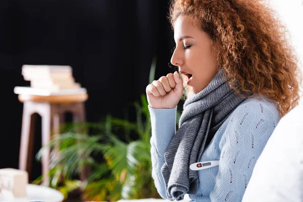 Selective focus of curly woman in scarf using thermometer and coughing at home — Stock Photo