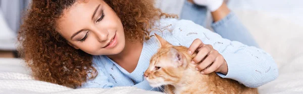 Orientação panorâmica da jovem mulher encaracolada acariciando gato tabby na cama — Fotografia de Stock
