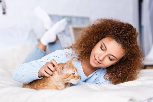 Focus selettivo della donna riccia che accarezza il gatto sul letto a casa — Foto stock