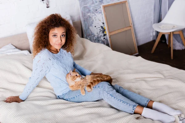 Femme aux cheveux rouges regardant la caméra près de chat tabby sur le lit — Photo de stock