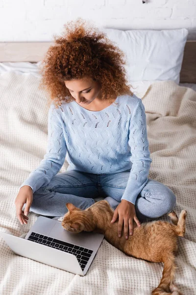 Donna dai capelli rossi accarezzando gatto vicino al computer portatile in camera da letto — Foto stock