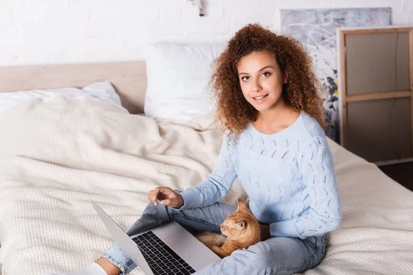 Рыжая женщина смотрит в камеру рядом с кошкой и ноутбуком на кровати — стоковое фото