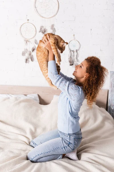 Vista lateral de una joven mujer rizada sosteniendo un gato pelirrojo mientras está sentada en la cama - foto de stock