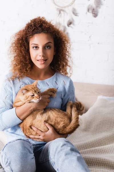Enfoque selectivo de la mujer joven mirando a la cámara mientras sostiene gato tabby en la cama - foto de stock
