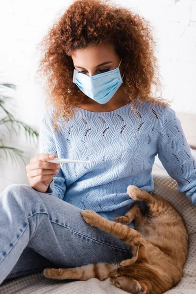 Foco seletivo da jovem mulher em máscara médica olhando para o termômetro ao lado do gato tabby na cama — Fotografia de Stock