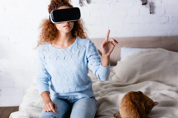 Mujer joven con auriculares vr apuntando con el dedo cerca del gato en el dormitorio - foto de stock