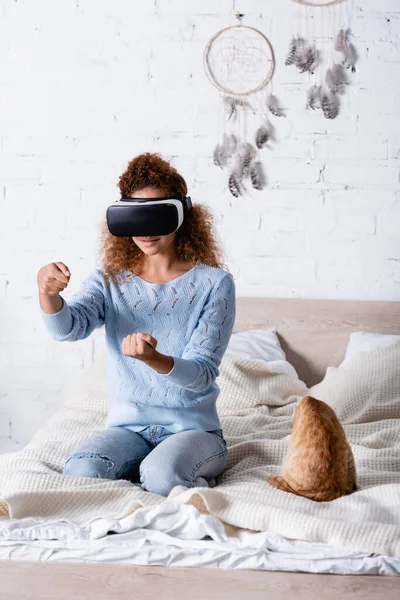 Focus selettivo della donna riccia utilizzando auricolare vr vicino al gatto in camera da letto — Foto stock