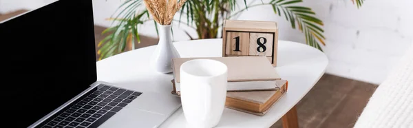 Panoramic shot of laptop with blank screen, books and cup on coffee table — Stock Photo