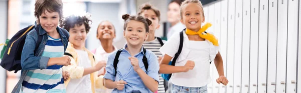 Панорамный снимок волнующих мультикультурных школьников, бегущих по школьному коридору — стоковое фото